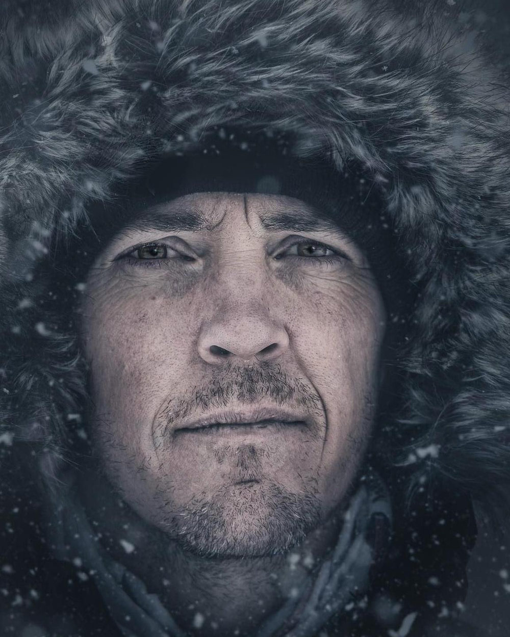 Bret Blakely in a fur-lined hood, snow falling, looking directly at the camera.